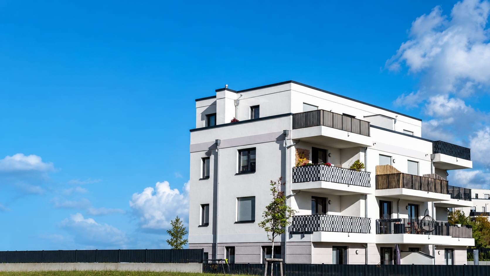 Edificio residencial moderno de viviendas con balcones y fachada blanca a la derecha, sobre un cielo azul despejado.